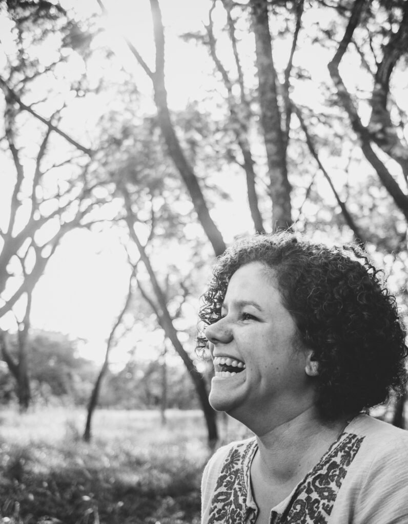 Fotografia em preto e branco da artista Maíra Freitas, mulher de pele clara e cabelos crespos escuro. A foto em preto e branco mostra seu rosto de perfil em uma grande risada, e parte de seu buscto com uma bata clara e ornamentos na gola. Ao fundo grama e árvores de troncos finos.